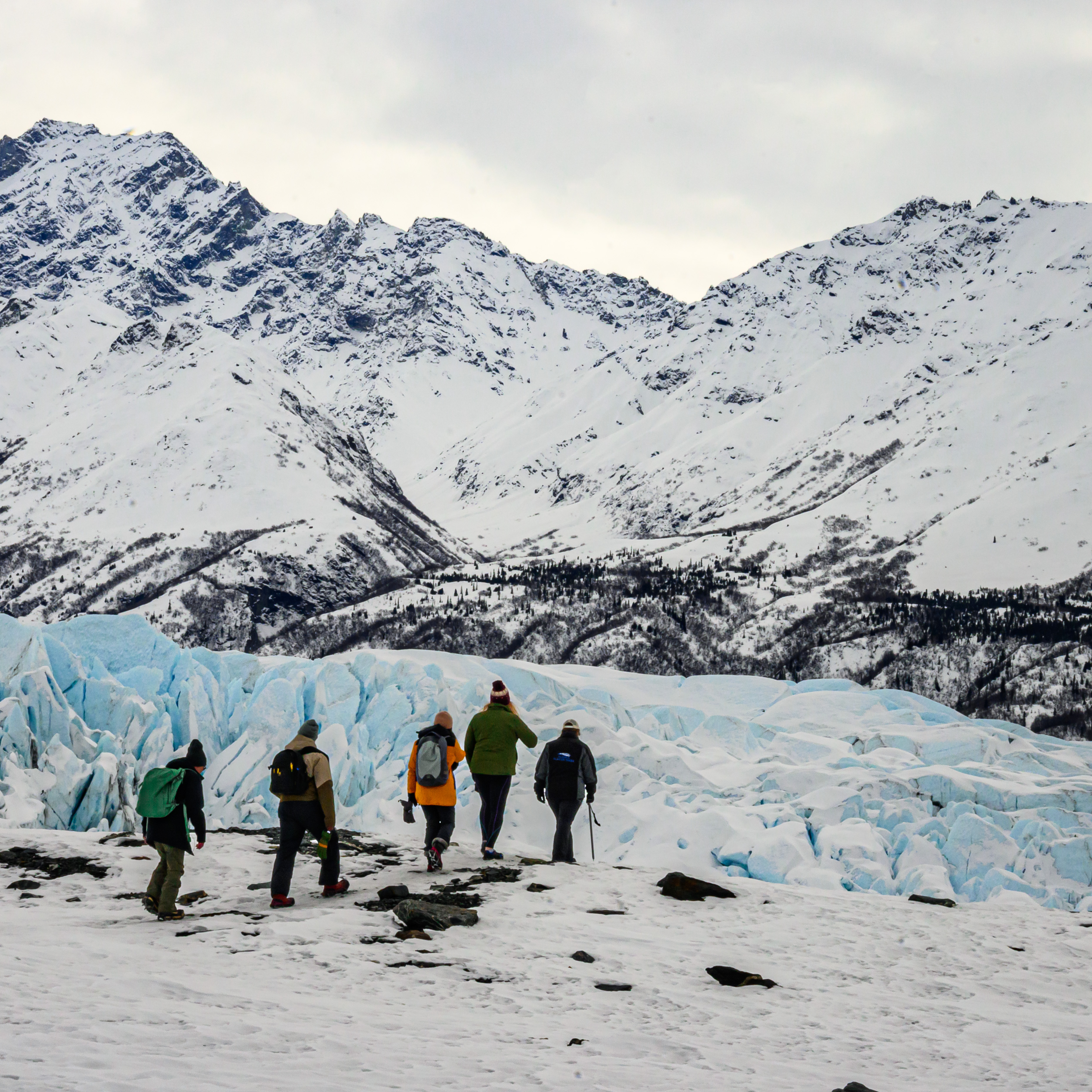 A group of hikers with backpacks walking across a snow and ice field approaching a massive blue glacier wall with snowy peaks behind